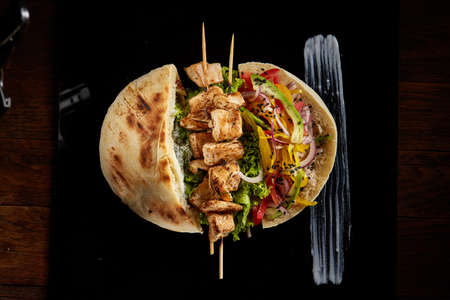 Tasty Jewish pita stuffed with grilled chicken, beans, vegetables, letucce and kebab with sauce on black square plate over vintage wooden background, top view, close-up. Restaurant serving. Traditional Middle East cuisine. Fast food concept.の写真素材