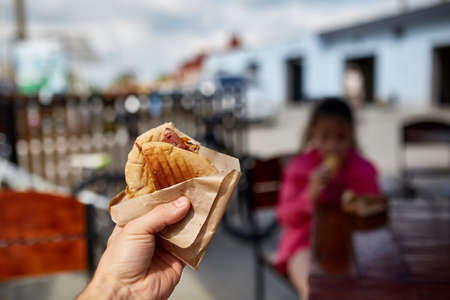 First person view shot of a man eating hamburger in the street cafe, soft focus, shallow depth of field. Yummy snack on the urban background. Delicious outdoor lunch in the open air cafe. Street food concept. Fast food concept.の写真素材