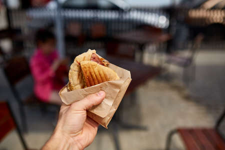 First person view shot of a man eating hamburger in the street cafe, soft focus, shallow depth of field. Yummy snack on the urban background. Delicious outdoor lunch in the open air cafe. Street food concept. Fast food concept.の写真素材