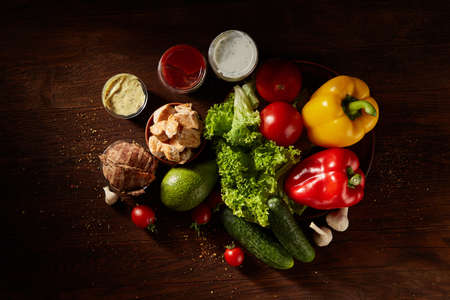 Juicy still life with pork chops, vegetables and sauces on a cutting board over a wooden background, top view, close-up, selective focus. Composition with fresh pepper, cucumber, squash, tomatoes, letucce. Barbecue background. Food concept.の写真素材