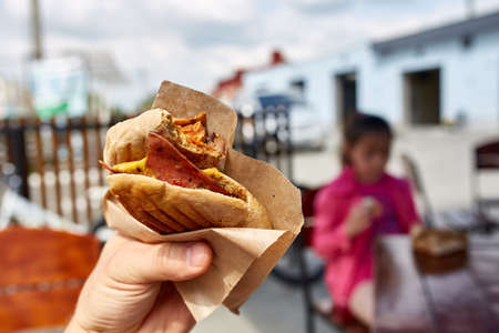 First person view shot of a man eating hamburger in the street cafe, soft focus, shallow depth of field. Yummy snack on the urban background. Delicious outdoor lunch in the open air cafe. Street food concept. Fast food concept.の写真素材