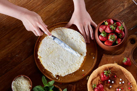 Close up of girl's hands adding some whipped sour cream on the top of delicious strawberry cake, selective focus. Tasty pastry is on the wooden table surrounded with organic ingredients. Homemade food concept. Baking with pleasure.の写真素材