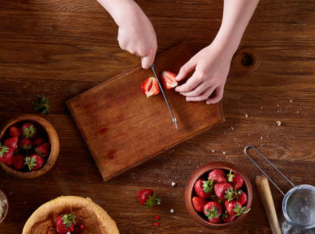 Close up of girl's hands adding some whipped sour cream on the top of delicious strawberry cake, selective focus. Tasty pastry is on the wooden table surrounded with organic ingredients. Homemade food concept. Baking with pleasure.の写真素材