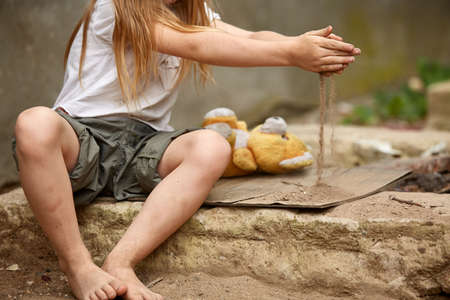 Orphan girl with bare feet in a shabby clothes playing with the dust on the street. Poverty and poorness posture. Lost childhood. Suffering children. Social inequality. Abandon generation. Poverty concept.の写真素材