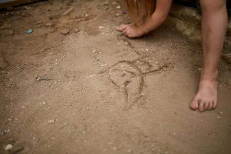 Lonely little girl drawing a cat with a pin in the sandy sidewalk, shallow depth of field. Street photo. Homeless orphan dreaming in the dirty street. Regret and sorrow about abandon children. Poverty concept.の写真素材