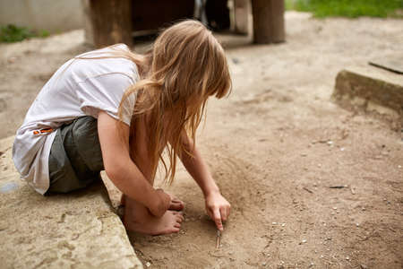 Concentrate lonely little girl drawing with a pin in the sandy sidewalk, selective focus. Street photo. Homeless orphan dreaming in the dirty street. Regret and sorrow about abandon children. Poverty concept.の写真素材