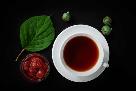 White cup with tea, strawberry jam, poppy boxes and hydrangea leaf on dark background, top view,...