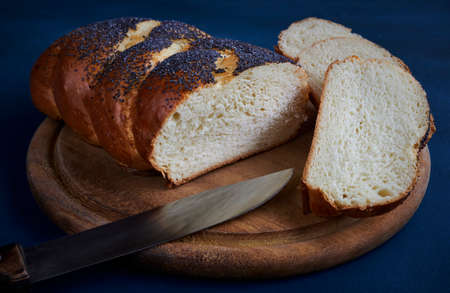 Fresh white bread loaf sprinkled with poppy on a chopping board with sliced slices and a knife, blue...