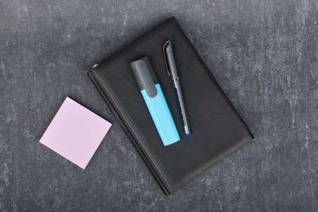 Ball pen and a marker lie on a black diary, next to paper sticks for pink records, business still...