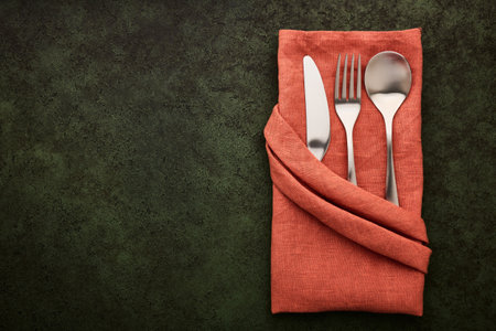 Terracotta-colored linen napkin folded in the shape of a pocket with cutlery inside, dark green background with copy spaceの写真素材