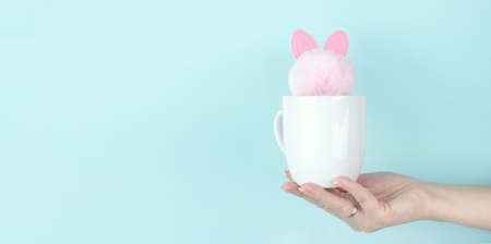Girl hand hold morning coffee cup with pink rabbit toy inside. Copy space. Blue backgroundの写真素材