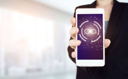 Infinite concept. Hand hold white smartphone with digital hologram infinity sign on light blurred...