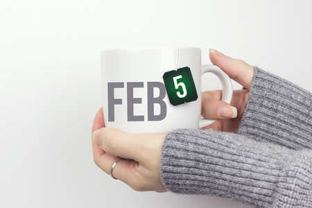 February 5th. Day 5 of month, Calendar date. Closeup of female hands in gray sweater holding cup of tea with month and calendar date on teabag label. Winter month, day of the year conceptの写真素材