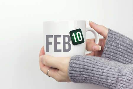 February 10th. Day 10 of month, Calendar date. Closeup of female hands in gray sweater holding cup of tea with month and calendar date on teabag label. Winter month, day of the year conceptの写真素材