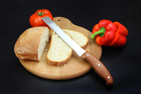 Freshly baked Bread, sliced with a serrated bread knife on a wooden cutting board with tomato and pepper on black backgroundの写真素材