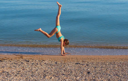 A young woman makes a wheel on the street. acrobatic gymnastics girl in a bikini on the beach. A young woman performing a pose at the water's edge. With space to copy.の写真素材