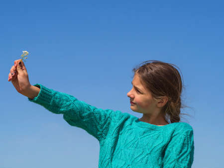 A girl with a flower in her outstretched hand against a blue sky background. A teenage girl with an outstretched arm illuminated by the sun on a blue sky background.の写真素材