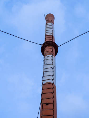 A large red and white non-working chimney with a ladder for climbing against a blue sky with white...