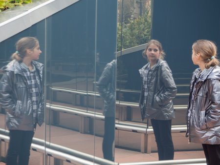 The girl is reflected on the mirror surface. A teenage girl in a gray jacket on vacation looks at a wall with a mirror surface that reflects her. A scene from real lifeの写真素材