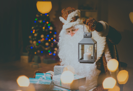 Santa brings and distributes gifts. Santa Claus with a candle lantern looking at gifts. . Cristmas tree.の写真素材