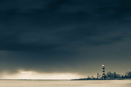 Dramatic cloudy sky over Baltic sea. Sea waves at long exposure. Striped lighthouse on the seashore. Toned black and white.の写真素材