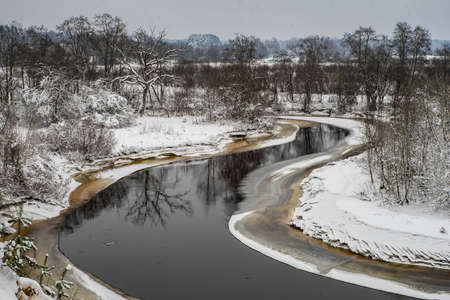 View of unfrozen river. Winter landscape. Snow on the ground and trees. Cloudy day.
