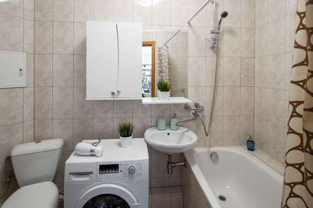 Simple interior of bathroom with beige tile. Bath, toilet and washing machine.