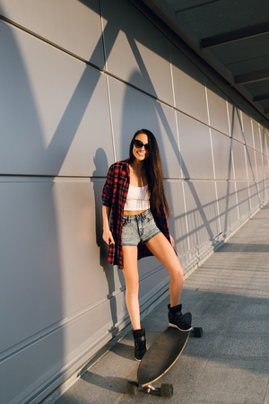 Pretty young fashion sexy swag girl riding wooden longboard on the road in evening summer time have fun and shows her nice back in jeans shorts hipster styleの写真素材