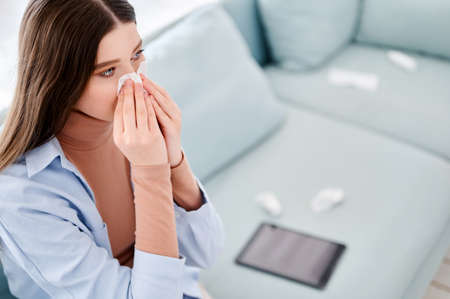 European young woman sneezing in tissue paper at home. Sick female sitting on sofa near tablet...