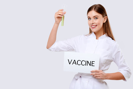 Young female doctor wearing medical coat holding tube of medicine and white card "Vaccine", smiling nurse on white isolated background, hospital workerの写真素材