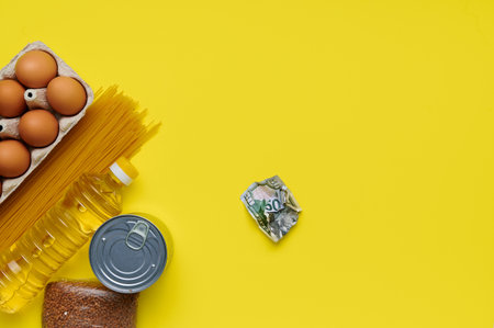 Chicken eggs, pasta, sunflower oil, buckwheat, canned food, paper bag, money, twisted us dollar. Food on yellow background, top view, flatlay. Pandemic help, cooking concept. Donation foodの写真素材