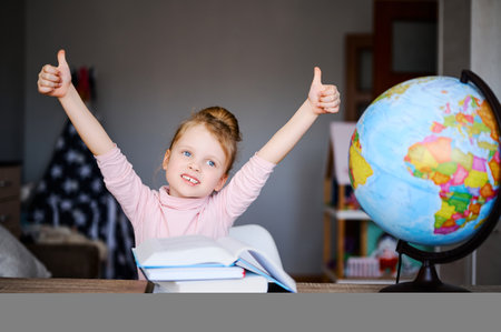 Little caucasian schoolgirl shows thumb up, reading books, doing homework. Training books, notebooks and globe at the table. Distance learning concept, online educationの写真素材