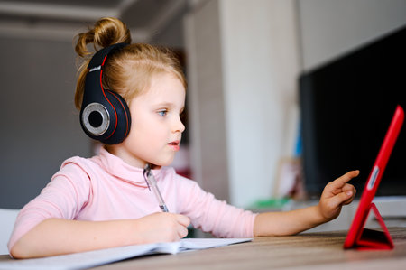 Distance learning online education. Schoolgirl in headphones studying at home with digital tablet and doing school homework. Training books and notebooks on tableの写真素材