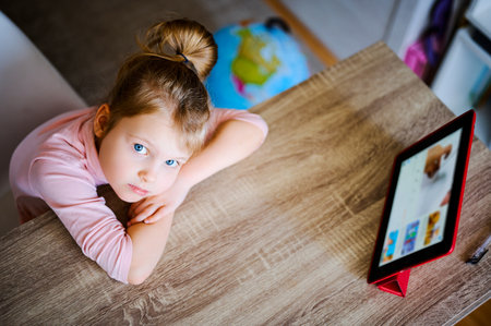 Young girl watches cartoons at home at a table on a digital tablet, a child plays with a device, a...
