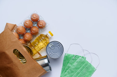 Food delivery, donation. Raw cereals, canned food, chicken eggs, sunflower oil, medical mask in paper bag on white background. Copy space, flat lay. Crisis food stock for coronavirus quarantineの写真素材