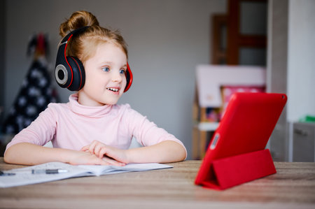 Cute little girl in headphones is using a tablet while doing homework and smiling, distance education, online learning, conceptの写真素材