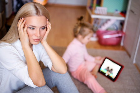 Young mother is nervous and sad at home on the floor, the child watches cartoons on the tablet and plays, naughty child, family concept.の写真素材