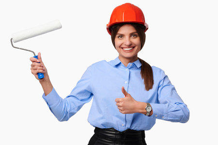 Smiling confident woman in helmet holding roller and showing thumb up ok gesture on white isolated backgroundの写真素材