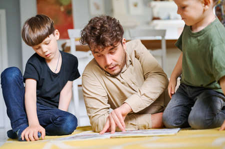 Father with sons reading book, spending time together at home and lying on the floorの写真素材