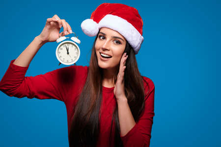 Eve celebration. Shouting caucasian woman wearing red santa fur hat holding clock, isolated blue backgroundの写真素材