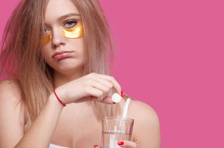 Sleepy young woman with patches under the eyes puts pain relieving tablet into glass of water. Girl after hangover early in the morning, pink isolated backgroundの写真素材