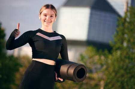 Smiling female holding yoga mat, showing thumb up gesture and listening music, city view, blurred...