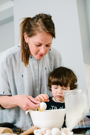 Mother young woman teaching showing her son child how breaking egg into a bowl.