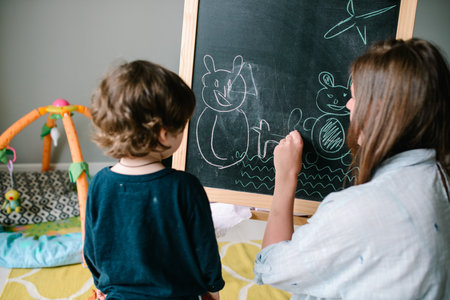 Young mother drawing chalk on blackboard with his young son at homeの写真素材