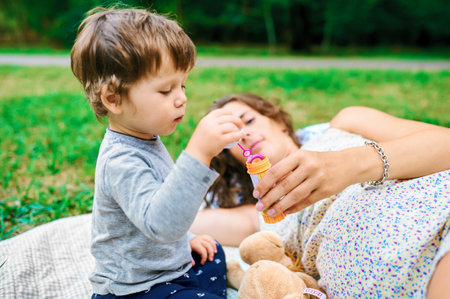 Young pregnant mother resting with her son in summer park and blow bubblesの写真素材