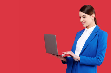 Attractive caucasian businesswoman wearing blue suit holding laptop and smiling on red isolated backgroundの写真素材