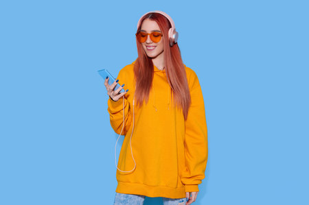 Young female in oversize hoodie listening to music on headphones and holding smartphone on bright...