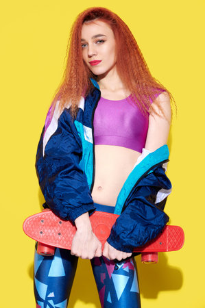Attractive hipster girl wearing bright colorful retro clothes holds skateboard on yellow background
