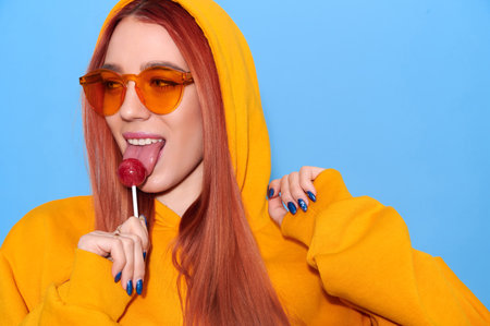 Attractive hipster girl wearing hoodie lick lollipop and show tongue on blue backgroundの写真素材