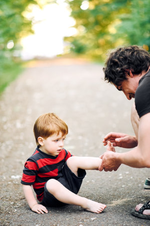 The relationship between son and father. Young father examines the leg of his young son in the summer in the parkの写真素材